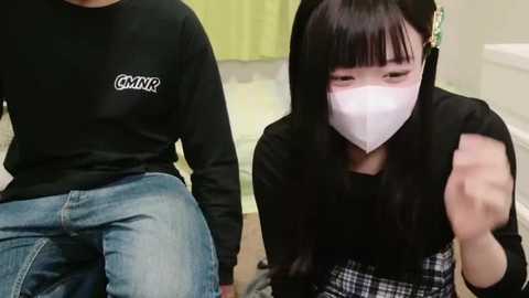 Media: Video of two Asian women in casual attire, one in a black long-sleeve shirt and blue jeans, the other in a black mask, checkered skirt, and green shirt, both indoors.