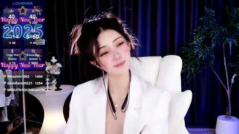 Media: Video of a fair-skinned Asian woman with dark hair in pigtails, wearing a white jacket, sitting in a white chair. Background includes a digital clock displaying 2025, a plant, and a white table with candles.