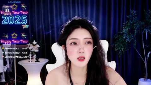 Media: Video of an East Asian woman with long black hair, wearing a black top, seated on a white chair. Background features a digital screen displaying \"2025\" and \"Happy New Year\" text, and a potted plant.