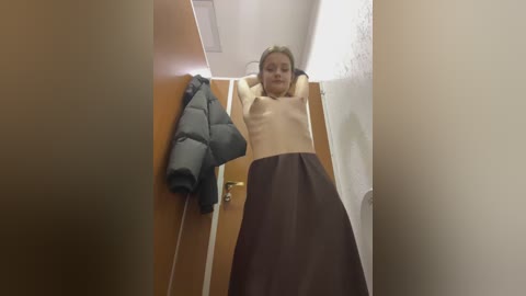 Media: Video of a young woman with light skin and brown hair, wearing a beige top and brown skirt, standing in a narrow, dimly lit hallway with a wooden door and a puffy jacket hanging on it.