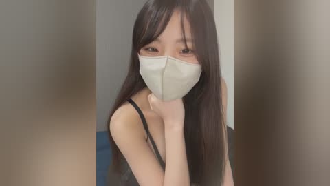 Media: Video of an East Asian woman with long black hair, wearing a white face mask and black tank top, leaning forward with her chin on her hand, in a dimly lit room with neutral walls.