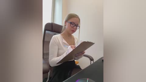 Media: Video of a blonde, fair-skinned woman with glasses, wearing a white cardigan and black skirt, seated at a desk, holding a clipboard and pen, in a modern, brightly lit office.