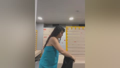 Media: Video of a woman with long, curly dark hair, wearing a turquoise towel, carrying a black bag, in a modern, brightly lit room with white and yellow storage units in the background.