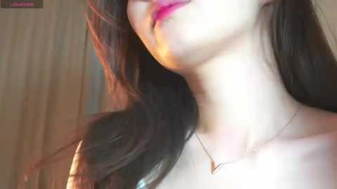 Media: A close-up video of a woman with long, dark hair and fair skin, wearing pink lipstick, a delicate necklace, and a soft, warm lighting that casts a glow on her face.