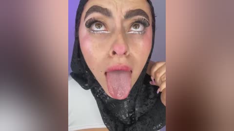 Media: Video of a young woman with light skin, large eyes, and dark eyebrows, wearing a black hijab, sticking out her tongue, with blurred faces in the background.