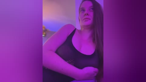 Media: Video of a young Caucasian woman with light skin, long brown hair, wearing a black tank top that reveals her breasts, standing in a dimly lit room with purple lighting.