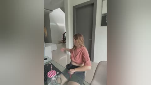 Media: Video of a young woman in a modern, minimalist apartment, playing a video game. She wears a pink crop top and black shorts, with a pink drink beside her.