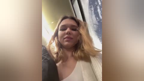 Media: Video of a blonde woman with shoulder-length hair, wearing a white top and a black jacket, standing in a train carriage with a blurred window showing snow.