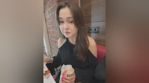 Media: Video of a young woman with fair skin and long brown hair, wearing a black off-the-shoulder top, sitting at a table, holding a white paper bag with a yellow logo. Brick wall and wooden paneling background.