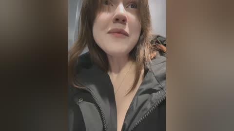 Media: A close-up video of a young woman with fair skin, shoulder-length brown hair, and a black jacket, partially unzipped, showing cleavage. She has a neutral expression.