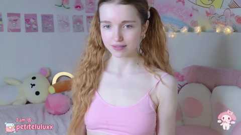 Media: Video of a young, fair-skinned, slender woman with long, wavy red hair in pigtails. She wears a pink spaghetti-strap tank top. Background features a soft pink, cartoon-themed room with plush toys and wall decorations.