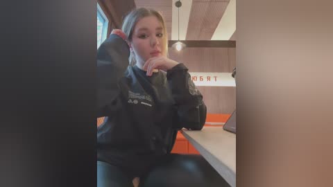 Media: Video of a blonde Caucasian woman with fair skin, wearing a black leather jacket, sitting at a counter in a modern, minimalist restaurant with orange seating and wooden decor.