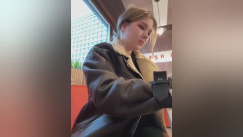 Media: Video of a young woman with blonde hair in a black leather jacket, fur-lined hood, and green pants, holding a black phone, standing in a modern interior with a large window and cityscape background.