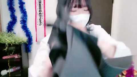 Media: Video of an Asian woman with dark hair and pale skin, wearing a white face mask, black shirt, and white gloves, sitting in an indoor room with blue tinsel, Japanese characters, and a green plant in the background.