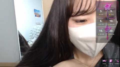 Media: A close-up video of an Asian woman with long, straight black hair and fair skin, wearing a white face mask, taken in a room with a bed and framed pictures on the wall.