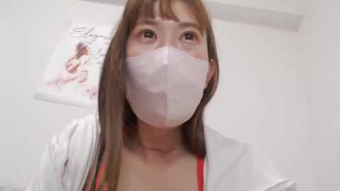 Media: A video of an Asian woman with long brown hair, wearing a white mask, white lab coat, and red top, standing indoors with a white wall and framed artwork in the background.