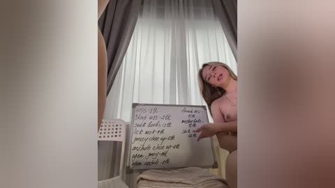 Media: A video of a nude, fair-skinned woman with long blonde hair, standing in a bedroom with a white sheet, gray curtains, and a chalkboard with handwritten text.