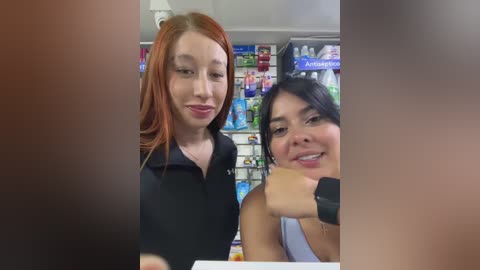 Media: Video of two women inside a convenience store. The woman on the left has red hair and wears a black hoodie, while the woman on the right has black hair, a sleeveless top, and a Fitbit on her wrist. The background shows shelves filled with products.