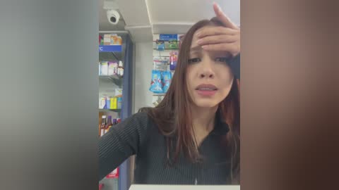 Media: A video of a young woman with long, straight brown hair, wearing a black sweater, wiping her forehead with her left hand in a dimly lit pharmacy aisle.