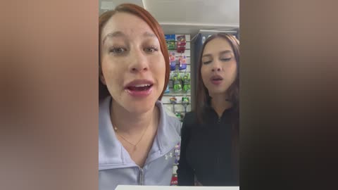 Media: Video of two women inside a convenience store. The woman on the left, with light skin and red hair, wears a light blue zip-up jacket. The woman on the right, with darker skin and long brown hair, wears a black top.