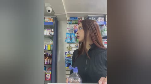 Media: A video of a woman with long, straight brown hair, wearing a black sweater, standing in a well-lit pharmacy aisle. She holds a plastic bottle of water and looks towards a shelf filled with various medicines.