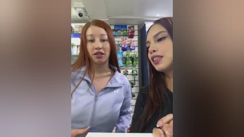 Media: Video of two women, one with long red hair, wearing a light blue zip-up, and another with long brown hair, wearing a black jacket, standing in a brightly lit, crowded store.