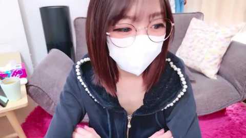 Media: Video of an Asian woman with glasses and a mask, wearing a black hoodie with pearl trim, sitting on a red carpet in a living room with a gray couch and colorful pillows.