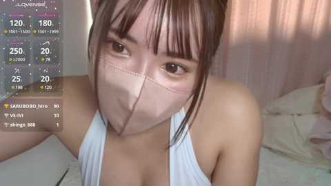 Media: Video of a young Asian woman with light skin, brown hair, and bangs, wearing a light blue tank top and a face mask, taken indoors with a pink curtain in the background.