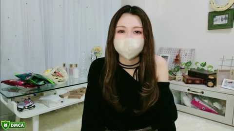 Media: A video of an Asian woman with long brown hair, wearing a black off-shoulder top, black choker, and a white face mask, standing in a brightly lit room with white walls, a glass table, and various decorative items.