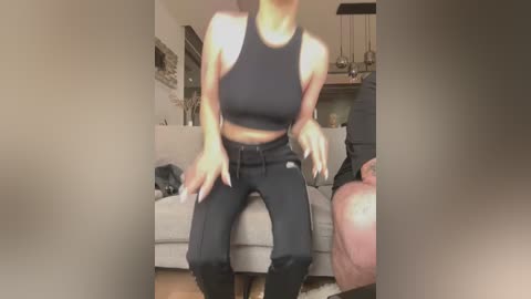 Media: Video of a woman with light skin, wearing a black sports bra and high-waisted black leggings, sitting on a beige sofa in a modern living room with a wooden floor.