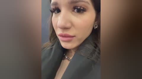 Media: A close-up video of a young woman with long dark hair, light skin, and full lips. She wears dark lipstick, a black jacket, and dangling earrings, set against a blurred, neutral background.