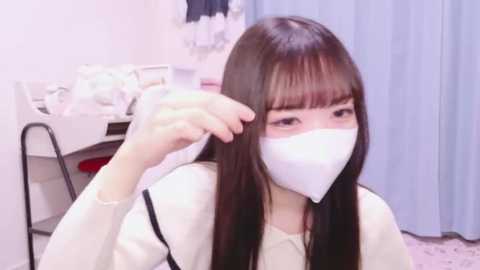 Media: Video of an East Asian woman with straight black hair, wearing a white mask, sitting in a room with light-colored walls, a white table, and blue curtains. She is adjusting her mask.