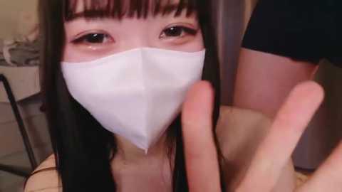 Media: Video of an East Asian woman with long black hair, wearing a white face mask, making a peace sign with her hands. She is indoors with blurred background.