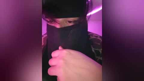 Media: A video of a person with dark hair and a black mask, partially obscured by shadows, holding a phone, under purple lighting.