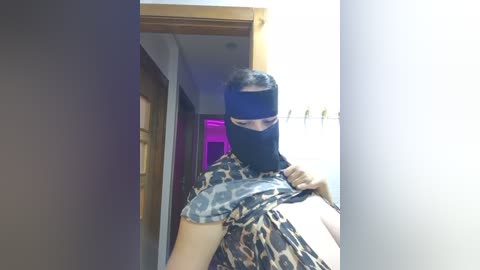 Media: Video of a person wearing a black balaclava, leopard print shirt, and holding a phone, standing in a dimly lit hallway with purple lighting.