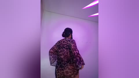 Media: A video of a person with dark hair in a bun, wearing a leopard print robe, standing in a minimalist white room with purple lighting, creating a dramatic, modern atmosphere.