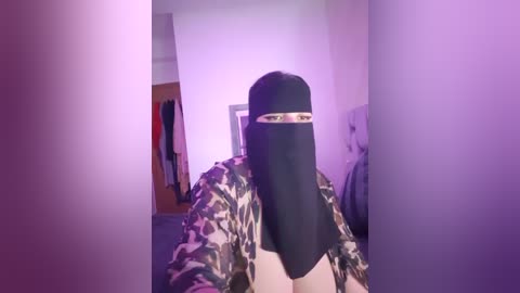 Media: Video of a person with long black hair, wearing a camouflage jacket, and a black niqab, standing in a dimly lit room with purple lighting. The room has a bed and a wooden wardrobe.