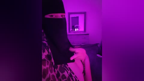 Media: A video with a purple tint shows a person with a leopard-print top, wearing a black face mask, touching their nose. The background features a framed mirror reflecting a room with dim lighting.