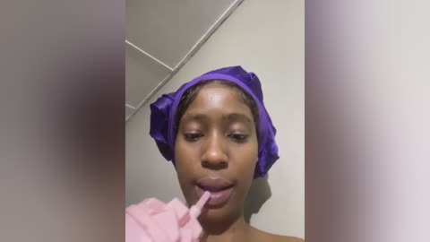 Media: Video of a young Black woman with a medium complexion, wearing a purple headwrap and brushing her teeth. The background features a beige wall and white ceiling tiles.