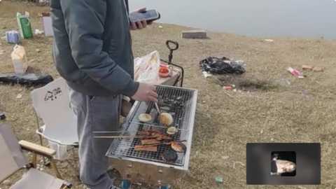 Media: Video of a person in a green jacket and jeans cooking hot dogs on a portable grill in a grassy, outdoor setting. The background includes scattered trash, a white chair, and a person holding a camera.