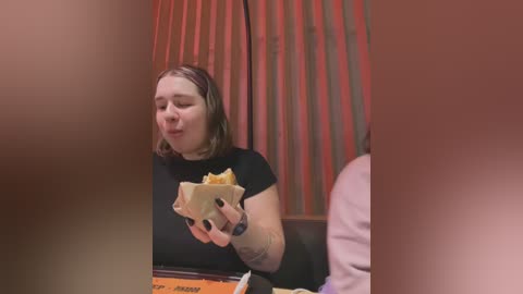 Media: Video of a woman with shoulder-length brown hair, wearing a black shirt, eating a burger, in a dimly lit restaurant with red curtains.