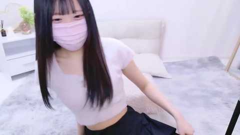 Media: Video of an East Asian woman with long black hair and fair skin, wearing a pink mask, white cropped top, and black skirt, sitting on a light grey carpet in a minimalist, white room.