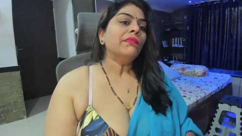 Media: Video of a middle-aged South Asian woman with medium skin tone, wearing a blue top and gold necklace, sitting on a bed in a dimly lit bedroom.