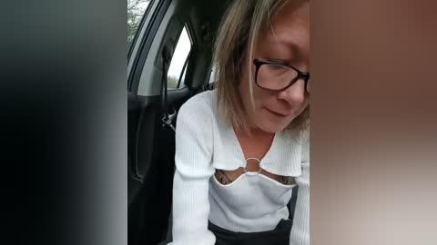 Media: Video of a middle-aged woman with glasses and light brown hair, wearing a white sweater and black pants, leaning against a car door, smiling slightly.