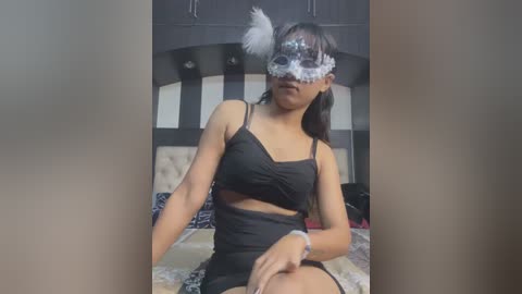 Media: Video of a young woman with medium skin tone, wearing a black dress with cutouts, feathered masquerade mask, and feathered headpiece, sitting on a bed with patterned bedding in a dimly lit bedroom.