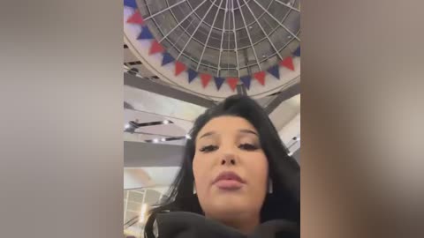 Media: A video of a young woman with long black hair, fair skin, and full lips, wearing a black jacket, in a modern, brightly-lit indoor setting with a circular ceiling and colorful pennant flags.