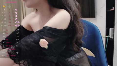 Media: Video of a woman with fair skin and long black hair, wearing a black lace top and a pink rose, seated in a blue chair. Background includes a calendar with social media icons and a white wall.