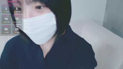 Media: A video of a person with a white surgical mask covering the mouth, wearing a black shirt, standing indoors with a blurred background and a digital clock overlay showing 2:15.