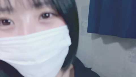 Media: A close-up video of an East Asian woman wearing a white surgical mask, covering her nose and mouth, with dark hair and brown eyes. She stands in front of a plain, light gray wall with a blue garment hanging nearby.