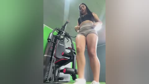 Media: Video of a curvy, light-skinned woman with long black hair in a gym, wearing a black crop top and gray shorts, standing on a treadmill.